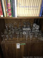 Wide assortment of clear glass stemware including wine, brandy, aperitif, and port glasses arranged on wooden shelf below books.