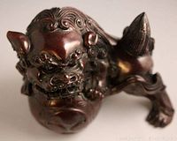 Angled close-up of Foo Dog figurine showing curled mane, fierce face, and paw on ball detail in burgundy red resin.