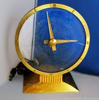 Frontal view of the brass and glass clock showing the clock face with brass numbers and hands, and attached black power cord.
