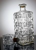 Front and side view of clear glass liquor decanter with textured square and floral geometric patterns, lying on its side showing the matching stopper with cork insert.
