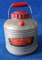 Front view photo of the vintage Knapp Monarch Therm-a-Jug metal thermos, showing red handle, screw lid, pour spout knob, and aged metal body with some wear and rust spots.