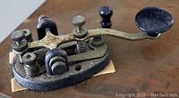 Close-up angled view of the metal vintage Morse code key showing the black knob, various adjustment screws and the rust on the metal surface mounted on plywood base.