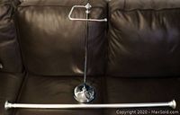 Photo showing the brushed chrome tension shower rod lying horizontally with rubber end caps visible on both ends and the weighted toilet paper holder stand upright.
