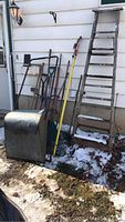 Photo showing metal snow pusher, various garden shovels, rakes, and an older wood step ladder leaning against a house.