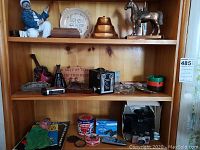 Three wooden shelves containing assorted figurines, cameras, ashtrays, and small collectibles including vintage cameras and decorative items.