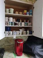 Open cabinet showing three shelves of assorted mugs, and red Keurig coffee machine below on counter