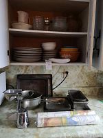 Shelves and counter with dishes, bowls, glassware, baking pans and metal grinder on countertop