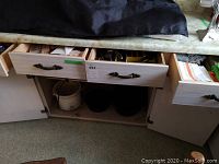 View of open drawers and cupboard showing silverware, utensils, and crock watcher inside cupboard.