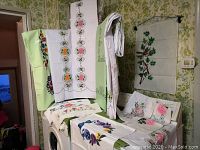 Multiple textile pieces including tablecloths, runners, and pillowcases displayed on and around a washing machine and hanging on walls. Fabric mostly white and green with floral designs.