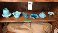 Overall view of six blue glassware pieces on a wooden shelf, showing variety of shapes and textures.