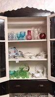 Full view of upper cabinet shelves showing blue goblets, red vase, clear floral pitcher, hand painted china plates with floral patterns, white milk glass bowls and vases, and green depression glass cups and small bowls.