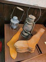 Two kerosene lanterns with 25 wooden sock forms on a wooden surface next to a wood-paneled wall.