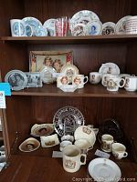 Three shelves of assorted commemorative china displayed in a wooden cabinet
