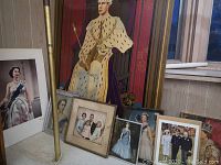 Seven framed pictures showing various royalty portraits. One prominent large colorful print of a king in coronation robes and crown sitting on an ornate throne. Smaller framed photos include queens in dresses and royal family group photos. Frames show wear and some damage.