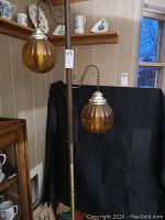 View of two tension rod pole lamps each with amber ribbed glass globes on curved metal arms.