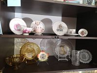 Two shelves of amber and clear glassware and porcelain items commemorating Queen Victoria's Jubilee including cups, saucers, plates, and bowls in amber and clear glass, with floral and royal motifs.
