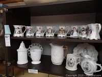 Two-tiered shelf with top shelf holding nine white porcelain scalloped mugs and pitchers with gold trim and royal portraits; bottom shelf holding milk glass stein, milk glass footed vase, white relief ceramic pitcher, black and white transfer mug and decorative plate