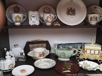 Two shelves display porcelain royal memorabilia including cups, plates, toiletry bottles, carriage figurine, and a decorative handbag.