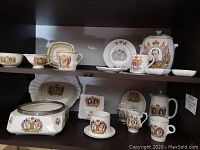 Porcelain collection on two shelf levels displaying cups, plates, pots, and jars bearing King George VI and Queen Elizabeth coronation portraits and heraldic motifs.