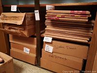 Eight boxes and several loose stacks of new oak wooden picture frames, size 12x16. Boxes are labeled as holding wood frames and 12x16 frames.