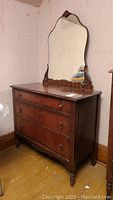 Full view of vintage wooden dresser with three drawers and scalloped-edge mirror on top, showing front and side angles with visible wear and ornate details.