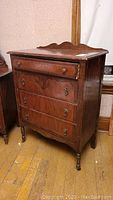 Front and angled view of the three-drawer dresser showing scalloped back panel, drawer knocker and pull hardware, and wood grain finish.