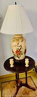Asian ceramic table lamp with cream ceramic base decorated with floral and bird images, topped with white pleated lampshade placed on round dark wood three-leg table along with two marble collectible vases