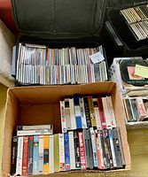Photo showing a cardboard box with VHS tapes and several plastic bins filled with VHS, CDs and cassettes, along with a black roller luggage containing CDs.