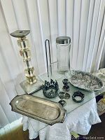 Wide shot of silver-tone and acrylic metal set showing candle holder, paper towel holder, glass container, tray, and metal decorative items on a covered table.