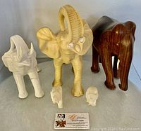 Front view of five elephant statues grouped on a table: three larger elephants in varied colors (ivory-colored, dark wood, and another ivory-colored), two small ivory or faux ivory miniatures in front.
