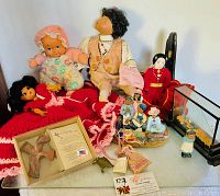 Overview photo of dolls and statues showing variety of items including dolls in various materials and styles, porcelain figurines, and a wooden cross with poem.