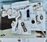 Photo of assorted vintage jewelry watches on white paper with notes, displaying various metal bracelet and wristwatch designs including rectangular, oval, square, and round dials.