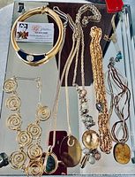 Top-down photo showing a variety of gold-tone costume jewelry necklaces, including coil design necklace with black enamel, spiral coil necklace with green pendant, and multiple chain necklaces with different pendants and bead accents.