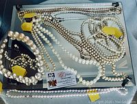 Multiple pearl necklaces and bracelets displayed on mirrored surface, showing variety of pearl sizes and strands with yellow tags.