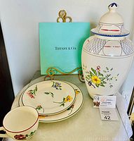 Small and large Tiffany & Co. plates and a coffee cup with floral designs in front of Tiffany & Co. signature green box, showing condition of plates and cup.