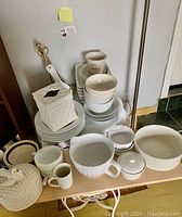 Multiple white and cream ceramic kitchenware and dishware items arranged on a table including plates, bowls, mugs, pitchers, and a textured lidded container with a basket weave design.