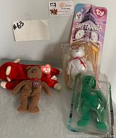 Photo of four Beanie Babies including a red teddy bear with Union Jack emblem and ty heart tag, Britannia the Bear in packaging, white bear, and green bear in packaging.
