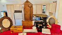 Overview of the lot including carved wooden frames, jewelry boxes in various sizes and styles, a large woven wicker container, a white ceramic decorative item, and a rectangular metal tray on a red surface with kitchen in background.