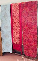 Three fabric runners hung over a chair, showing decorative patterns in blue with gold, red with tan, and dark red floral designs.