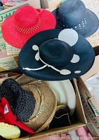 Photo showing red woven sunhat, two black broad-brim hats with white dots and white decorative abstract design, and a box containing straw woven hats and other pieces.