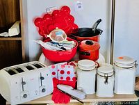 Photo showing a collection of red and white kitchenware including a white toaster, ceramic canisters, red pots, black skillet, oven gloves, mixing bowl with utensils, and trays arranged on a surface.