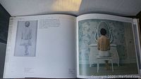 Open pages of the book showing one painting of a woman sitting in front of a mirror in a detailed interior setting, including text and a smaller illustrative drawing on the left page.