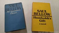 Photo showing two hardcover books by Saul Bellow: Mr. Sammler's Planet with blue cover and Humboldt's Gift with yellow cover placed side by side on a beige surface.