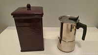 Photo showing side view of maroon ceramic biscotti cookie jar and stainless steel espresso coffee maker on a flat surface.