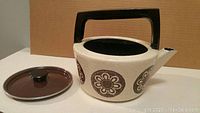 Side view of the beige enamel on steel tea kettle with floral medallions and black rectangular handle, lid beside it.