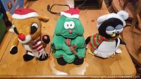 Front view of three plush holiday decorations: Rudolph the Red-Nosed Reindeer, Christmas tree, and a penguin, displayed on a wooden surface.