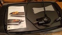Top view of Wolfgang Puck electric reversible grill showing large nonstick cooking surface, temperature control dial, power cord, and included plastic spatulas and user manual.