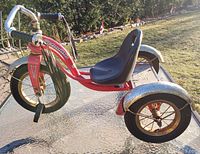 Side view of the red Schwinn Roadster tricycle showing the adjustable black seat, chrome handlebars with tassels, and steel fenders with some rust.