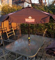 Black Sleeman branded patio umbrella open and set through a glass-top patio table, surrounded by metal chairs, showing the overall outdoor setup and fabric canopy with logos.