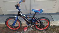 Blue Spiderman-themed child bike with red rims, red pedals, spider graphics on seat and frame, taken from side view against closed garage door